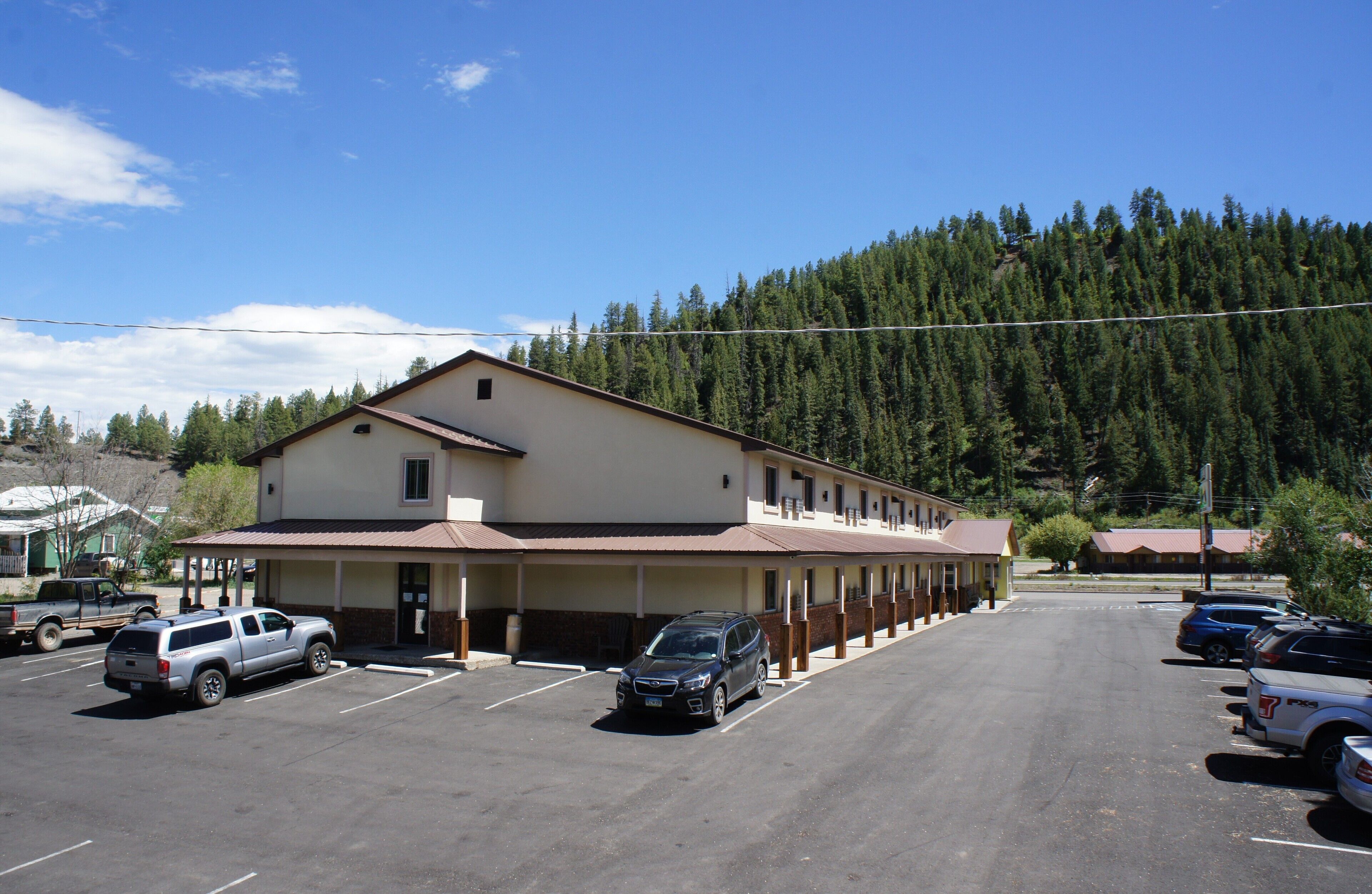 A motel with a covered parking area, a row of rooms, and a forested mountain backdrop.