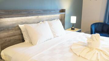 Habitación de hotel con cabecera de madera, ropa de cama blanca, un arreglo de toallas, una mesita de noche con lámpara y una silla azul.