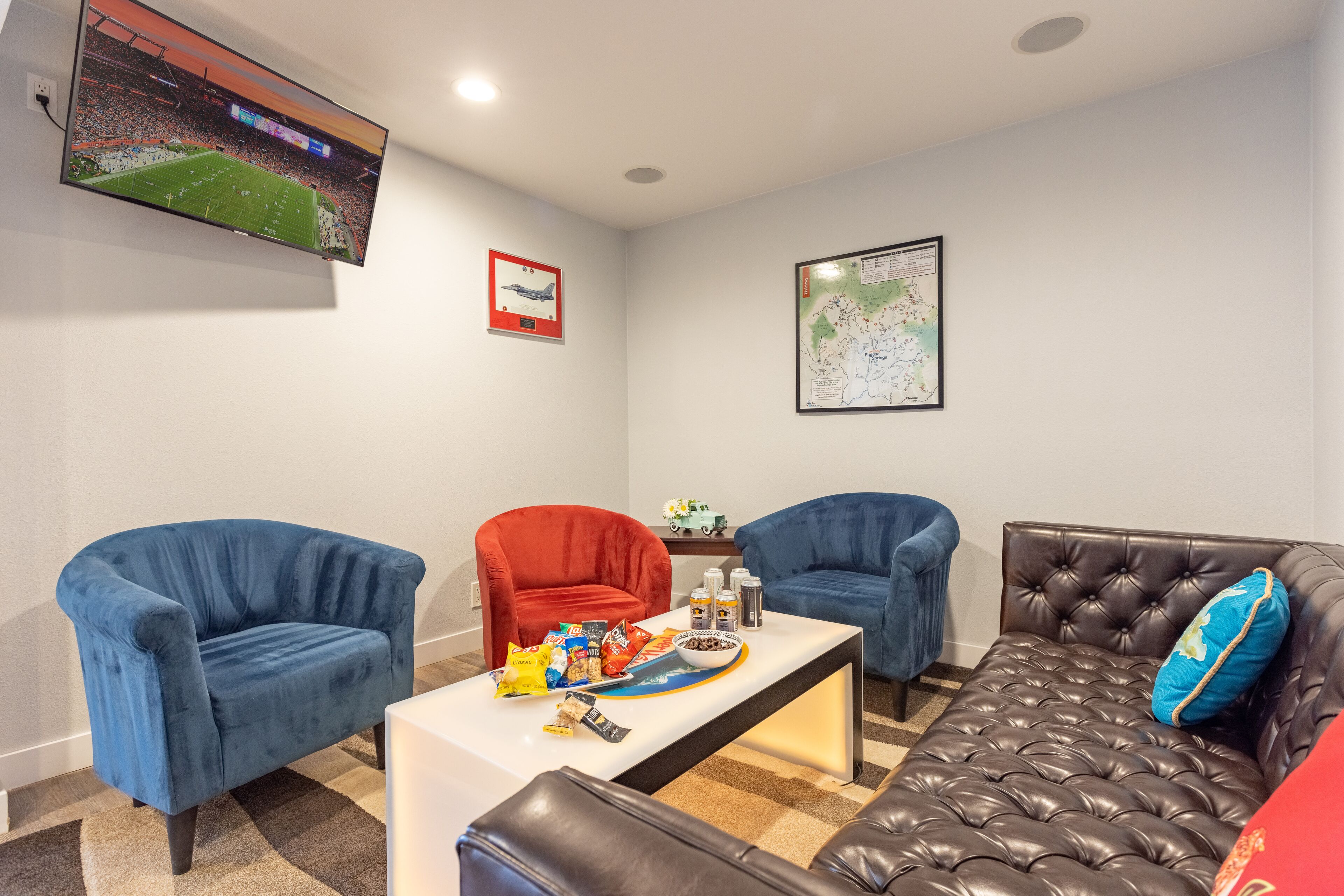 A living room with a flat-screen TV, two blue armchairs, a red armchair, a brown leather sofa, and a coffee table with snacks.