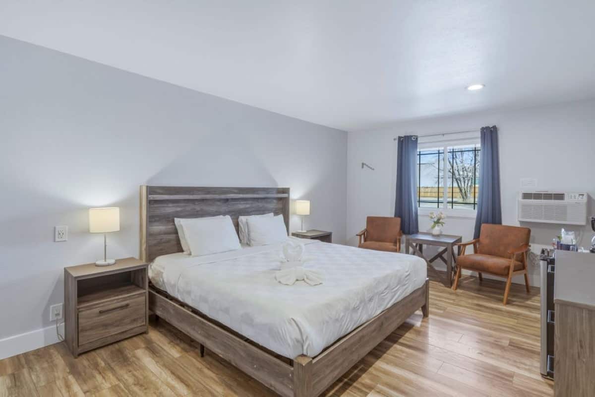 A bedroom with a wooden bed, bedside tables, a desk, two chairs, a window with curtains, and an air conditioning unit.