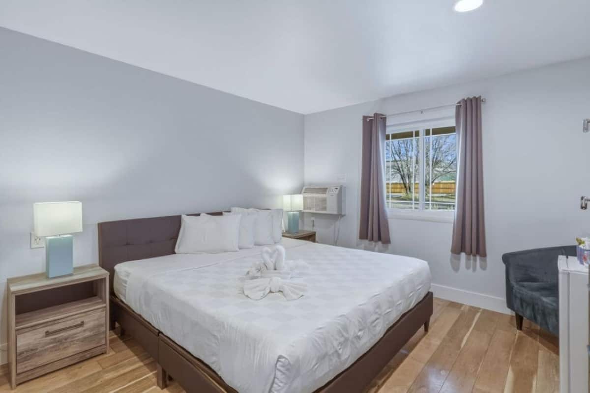 A hotel room with a bed, bedside tables, a desk, a window with curtains, and an air conditioning unit.