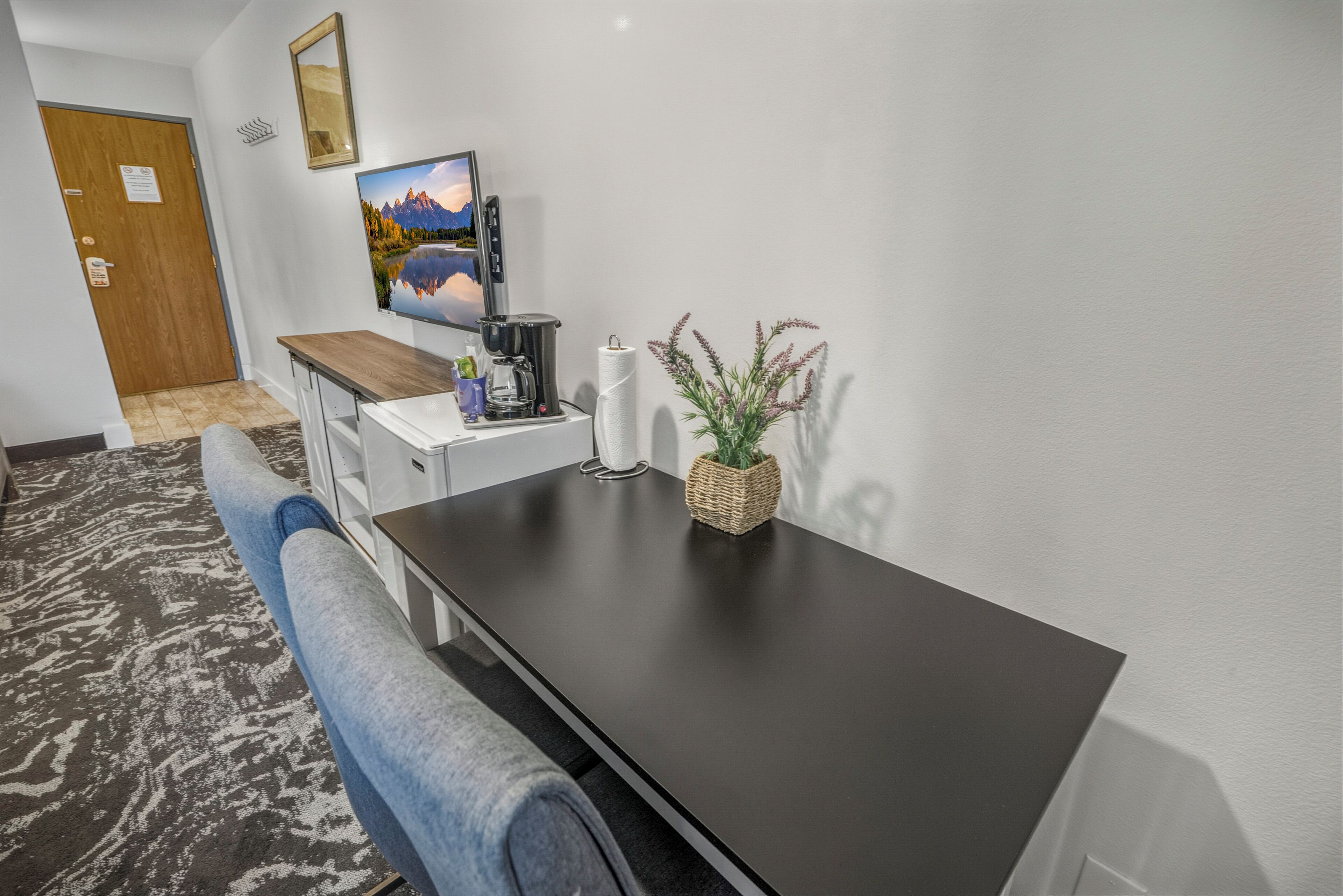 A hotel room with a dark wooden desk, a coffee maker, a TV, and a plant on the wall.