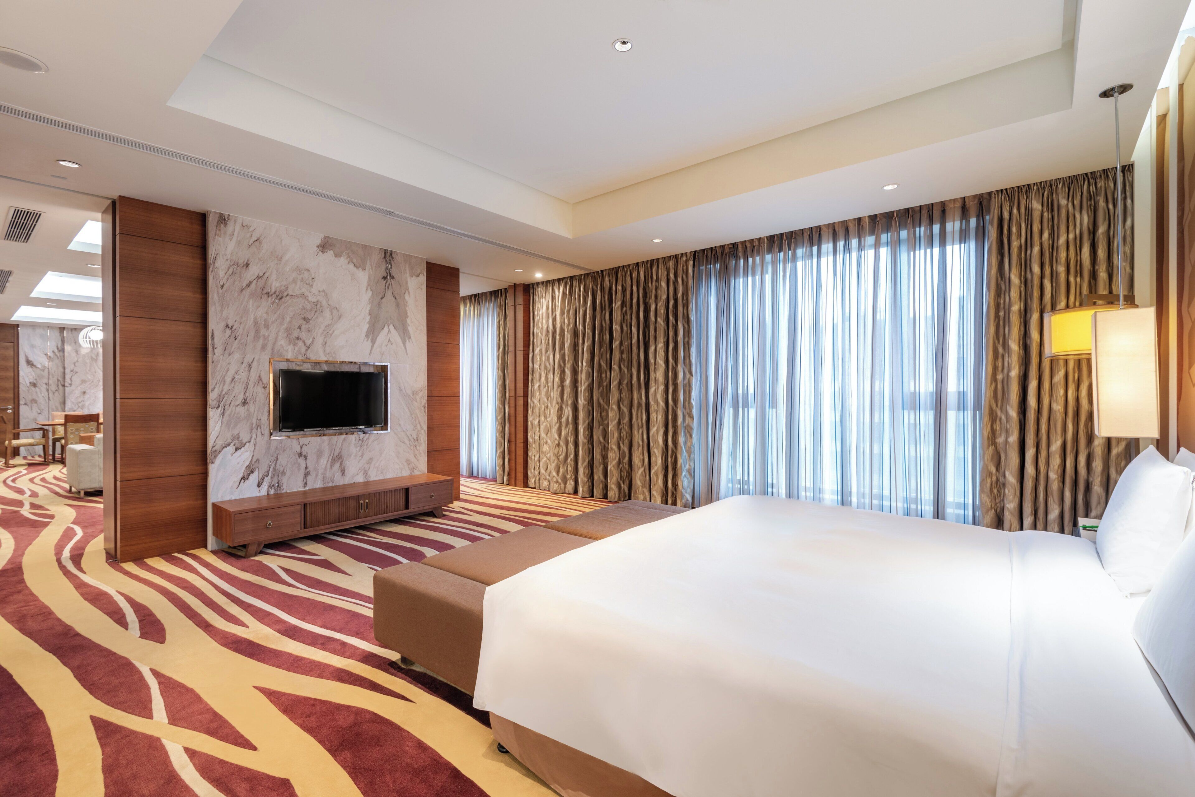 A hotel room with a large bed, a flat-screen TV mounted on a marble wall, a sofa, and a patterned carpet.