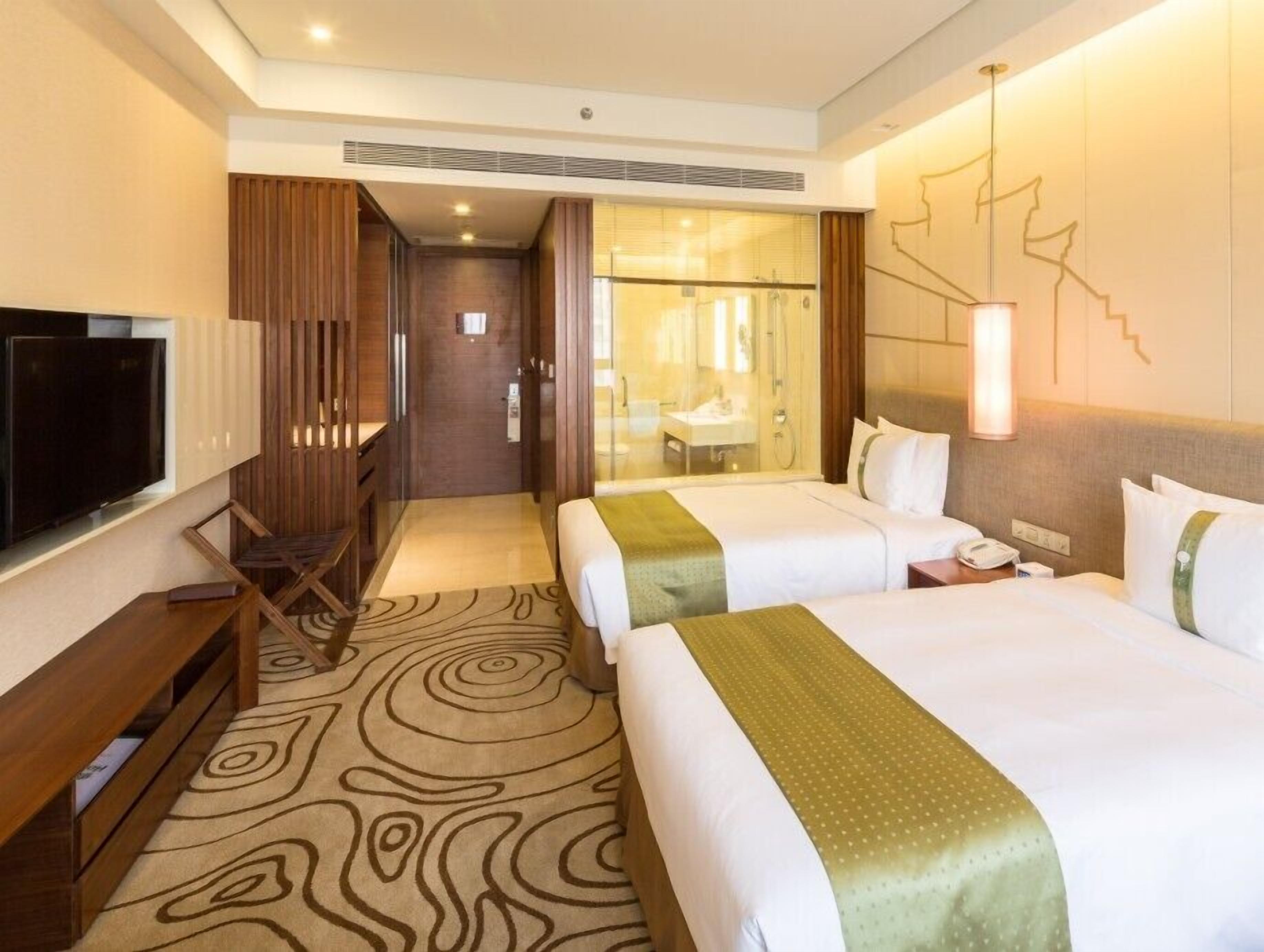 A hotel room with two beds, a TV, a desk, and a bathroom visible through a glass door.