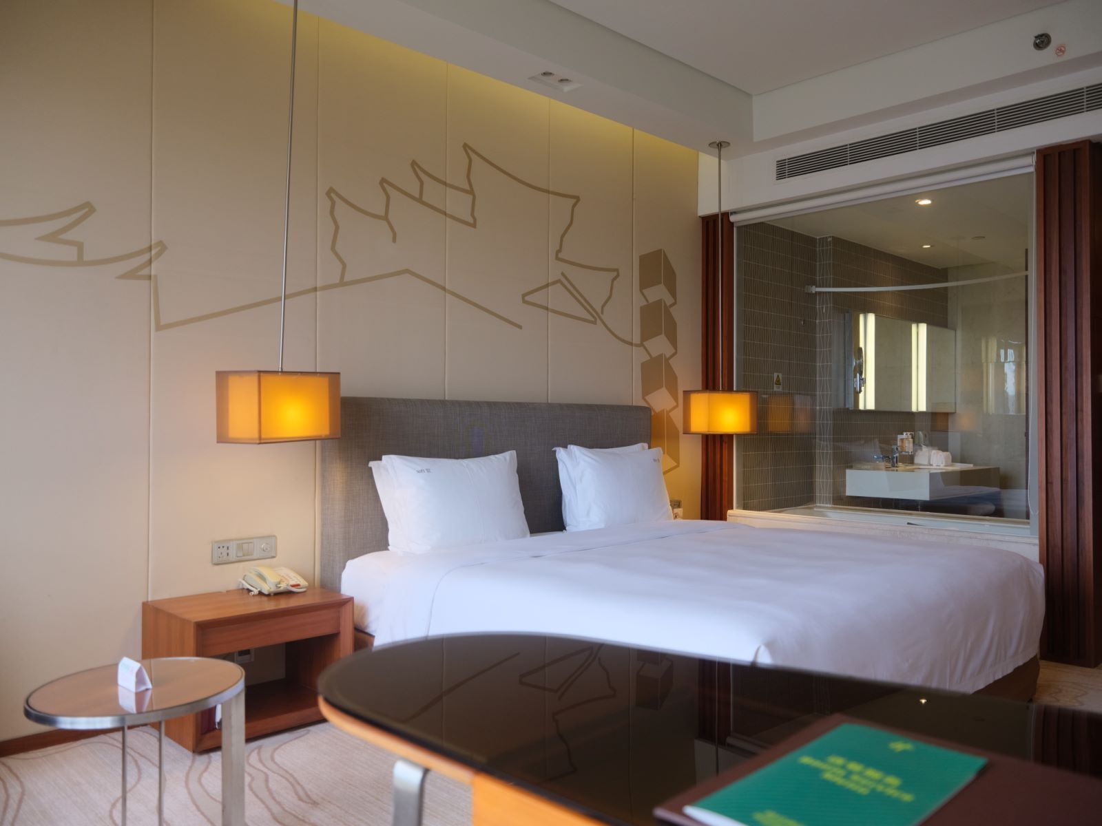 A hotel room with a large bed, bedside lamps, a small table, a mirror, and a shower area visible through a glass door.