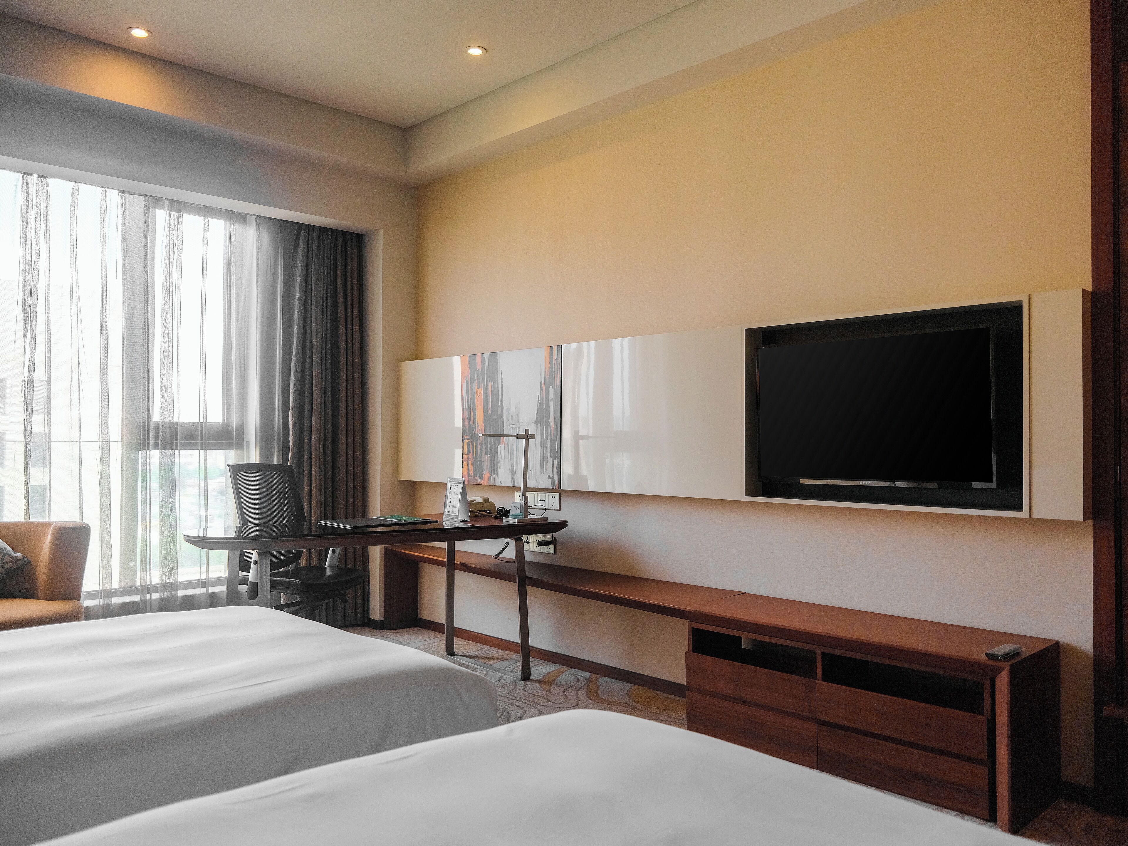 A hotel room with two beds, a desk with a chair, a flat-screen TV mounted on the wall, and a large window with curtains.