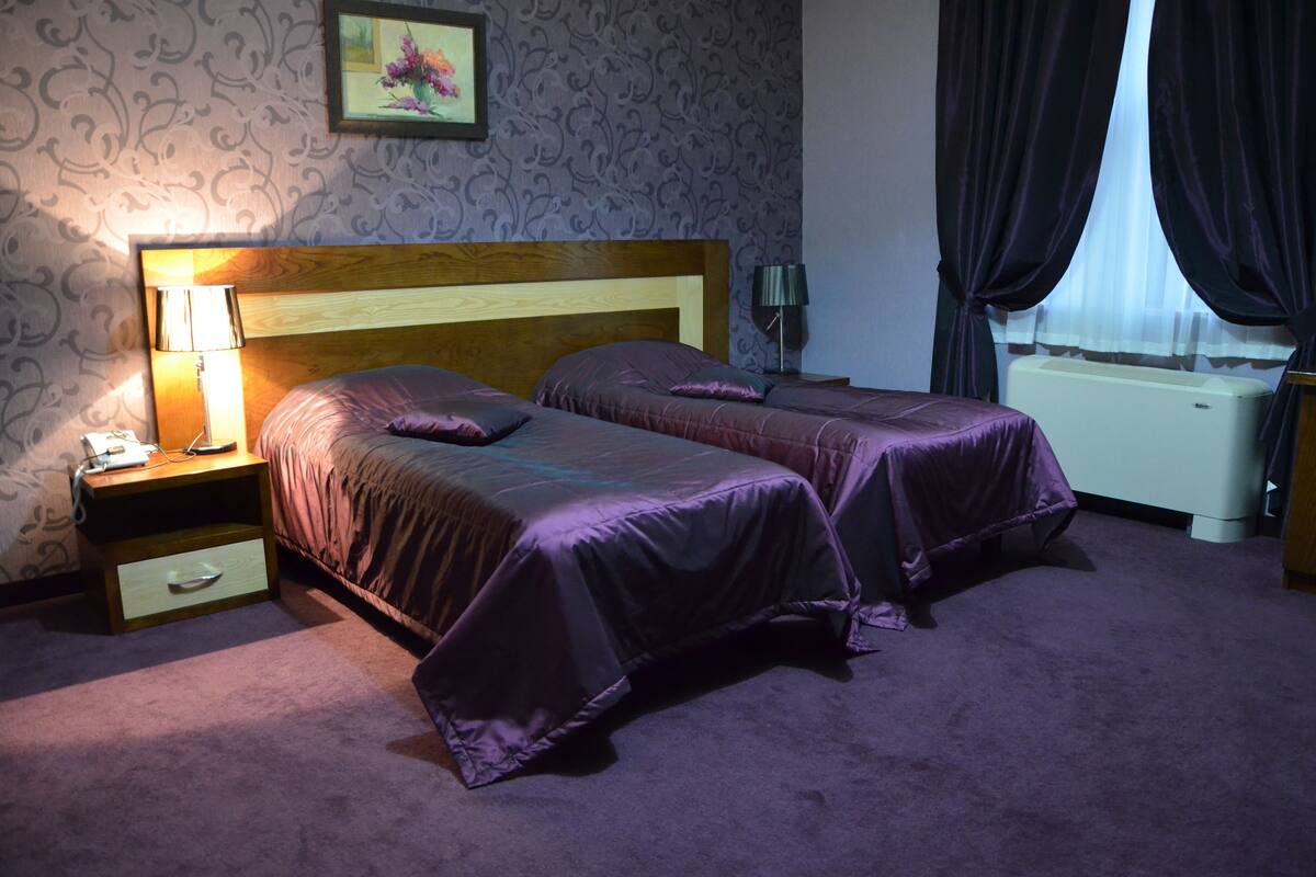 A hotel room with two beds, a nightstand with a lamp, a wall clock, and a radiator.