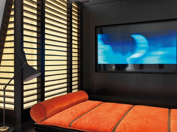 Una habitación moderna con un televisor con marco negro en la pared, una lámpara de pie negra y un banco con cojín rojo.