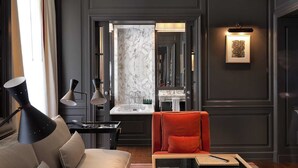 A modern living room with a dark wood-paneled wall, a marble fireplace, a sofa with cushions, a red armchair, and a side table with a lamp. - Marquis Faubourg Saint Honoré - Small Luxury hotels of the World (Paris)