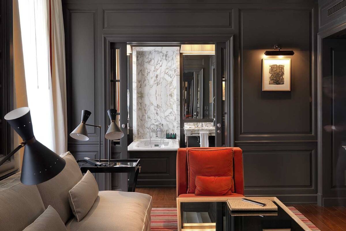 Un salon moderne avec un mur lambrissé en bois foncé, une cheminée en marbre, un canapé avec des coussins, un fauteuil rouge et une table d’appoint avec une lampe.