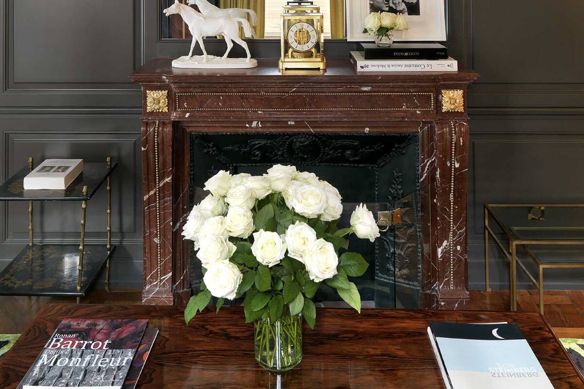 Une pièce avec une cheminée en bois sombre, un vase blanc rempli de fleurs et une cheminée ornée d’objets décoratifs.
