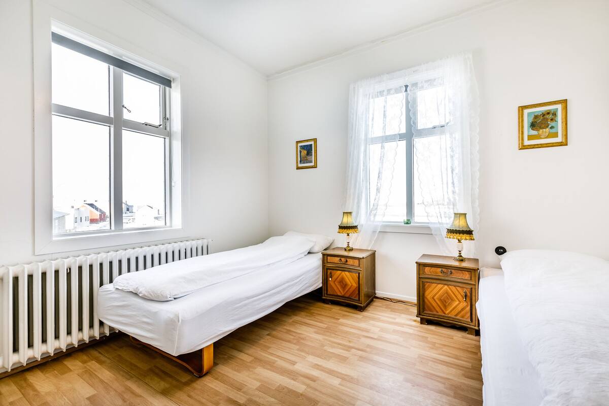 Ett sovrum med en enkelsäng, ett träbord vid sängen med en lampa, ett element och två fönster med gardiner.