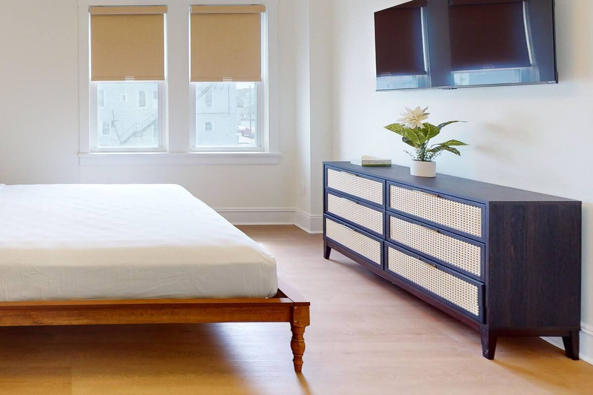 A bedroom with a bed, a dresser, a TV, and two windows with blinds.