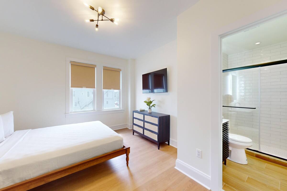 A modern bedroom with a wooden bed, a dark dresser, a flat-screen TV, and a bathroom with a glass shower enclosure.