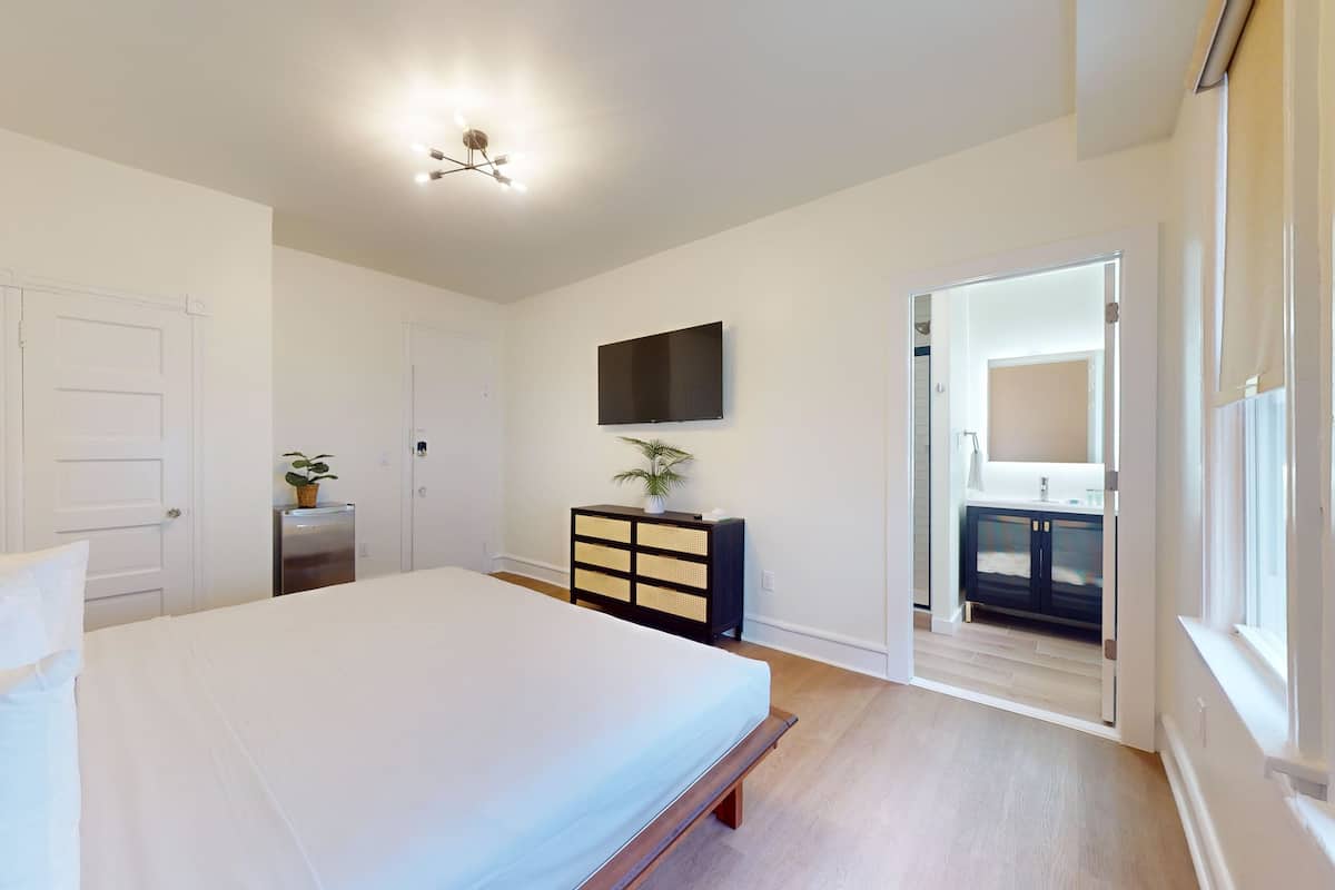 A bedroom with a bed, a dresser, a TV, and a door leading to a bathroom.