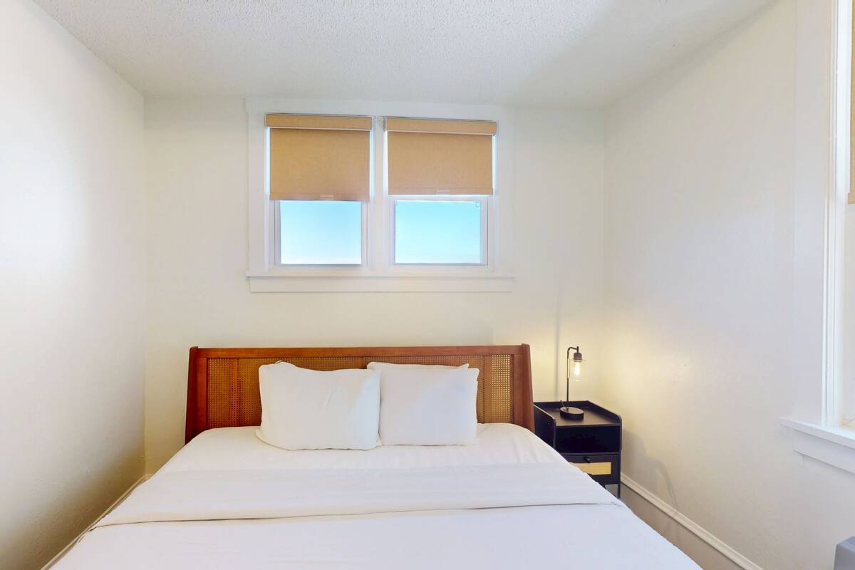 A small, clean room with a single bed, a nightstand, a lamp, and a window with blinds.