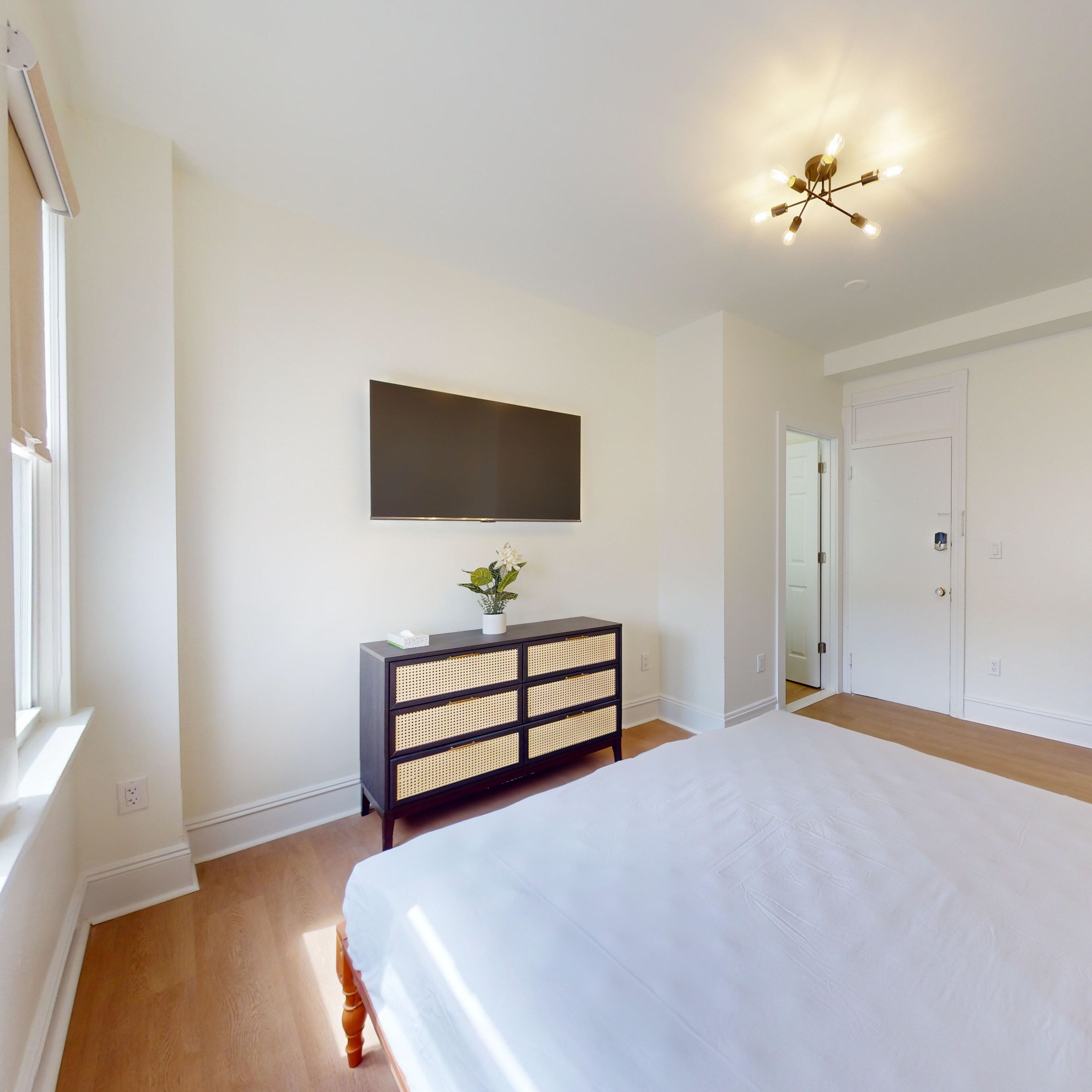 Ein Schlafzimmer mit einem Bett, einer Kommode, einem an der Wand befestigten Fernseher und einer Tür.