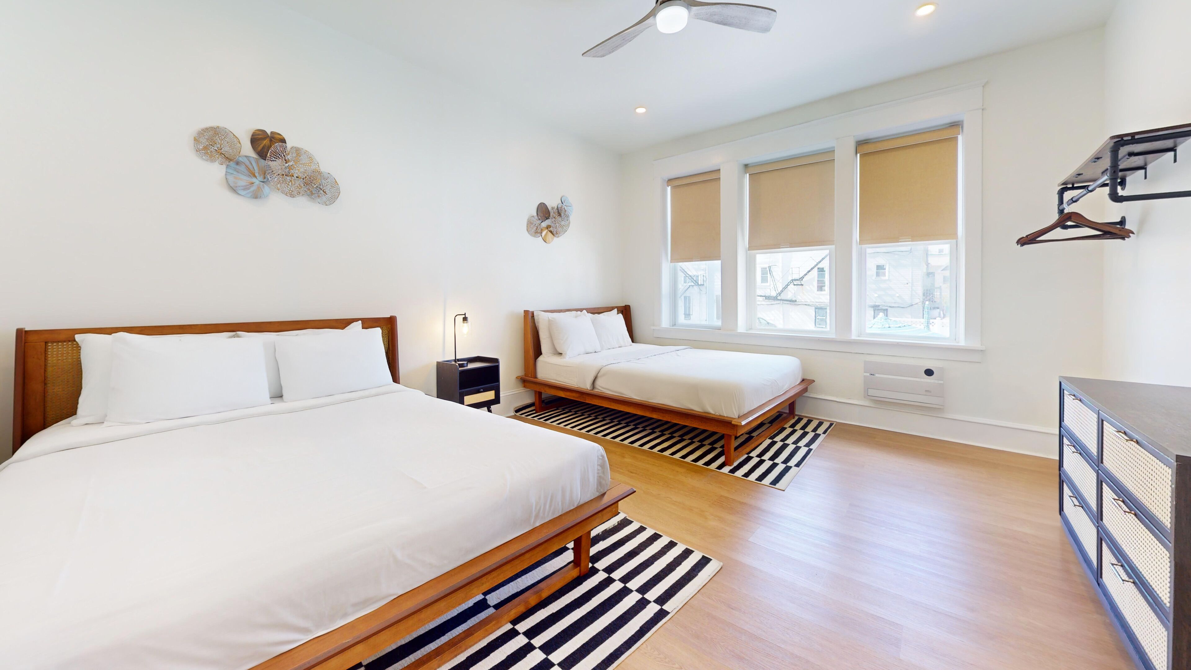A modern hotel room with two beds, a ceiling fan, large windows, and a view of the city.