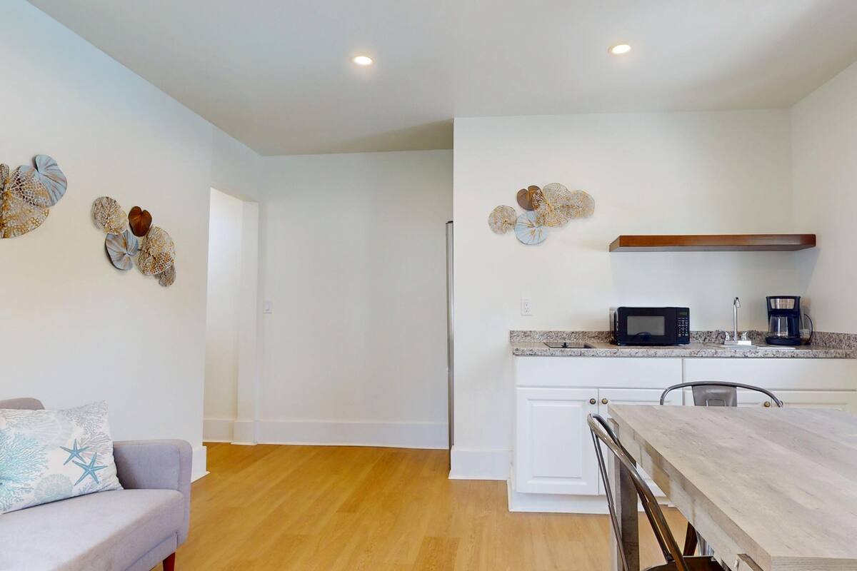 A modern kitchen with white cabinets, a microwave, and a coffee maker. There is a dining table with chairs and a sofa with a decorative pillow.