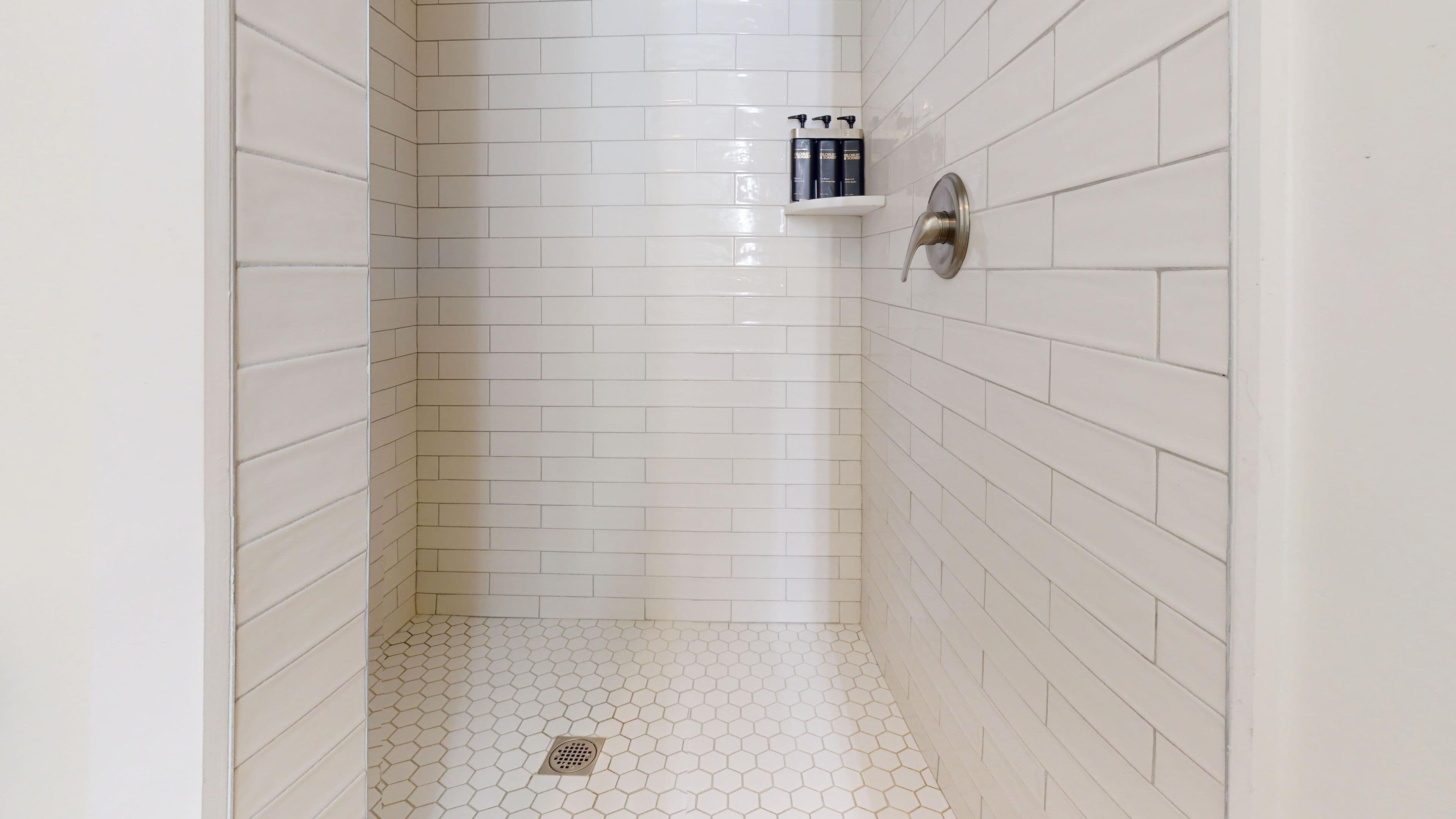 A shower with white hexagonal tiles, a silver showerhead, and a shelf with toiletries.