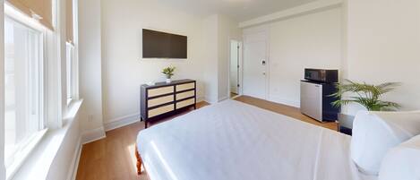 A bedroom with a bed, a dresser, a television, a refrigerator, and a potted plant.