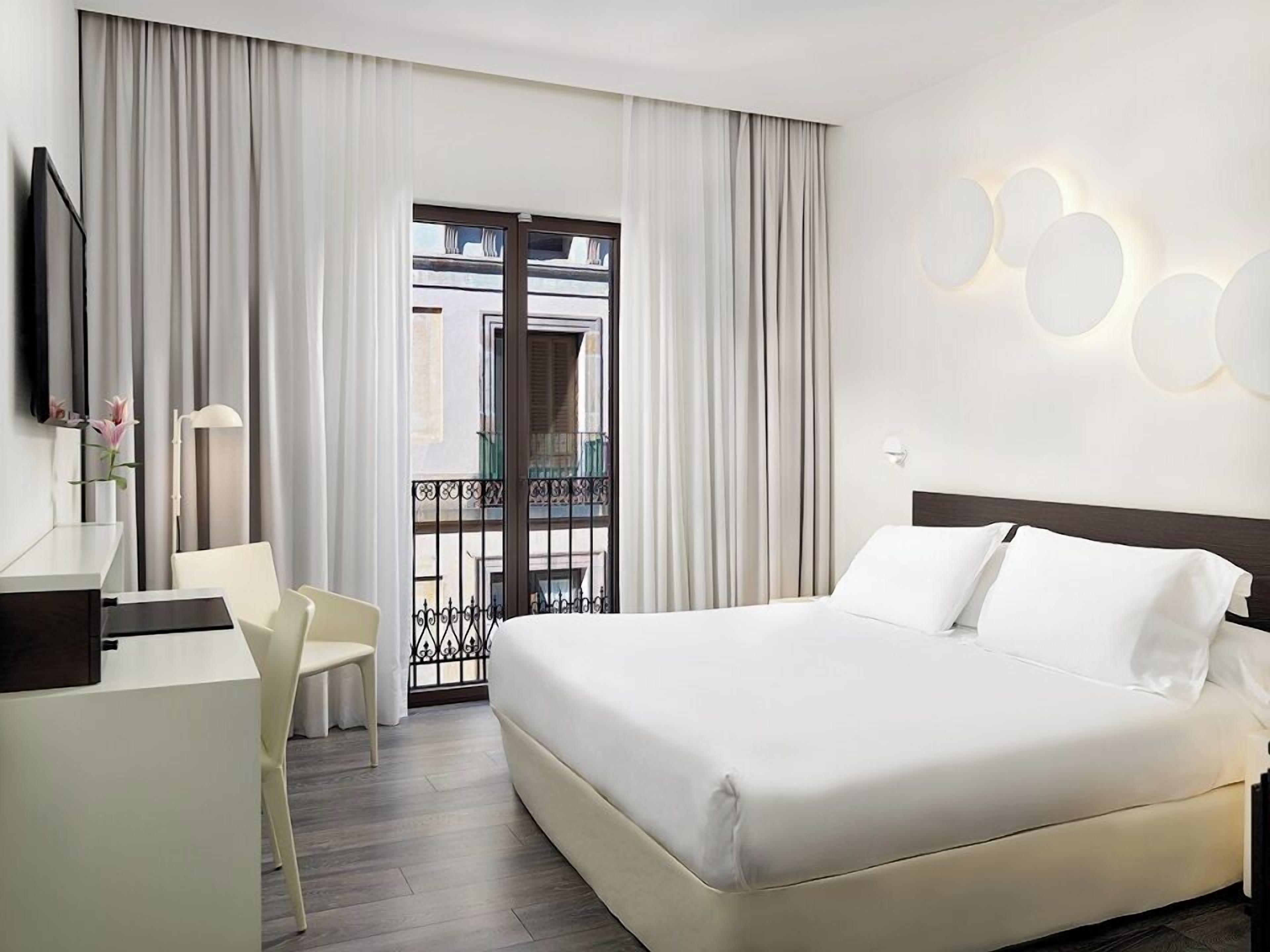 Quarto de hotel com cama, escrivaninha, cadeira e varanda com cortinas.