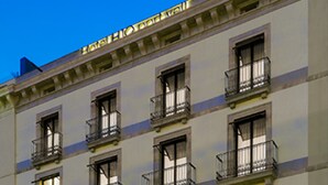 Un hôtel à plusieurs étages, doté de balcons, de boutiques au rez-de-chaussée et d’une façade bien éclairée.