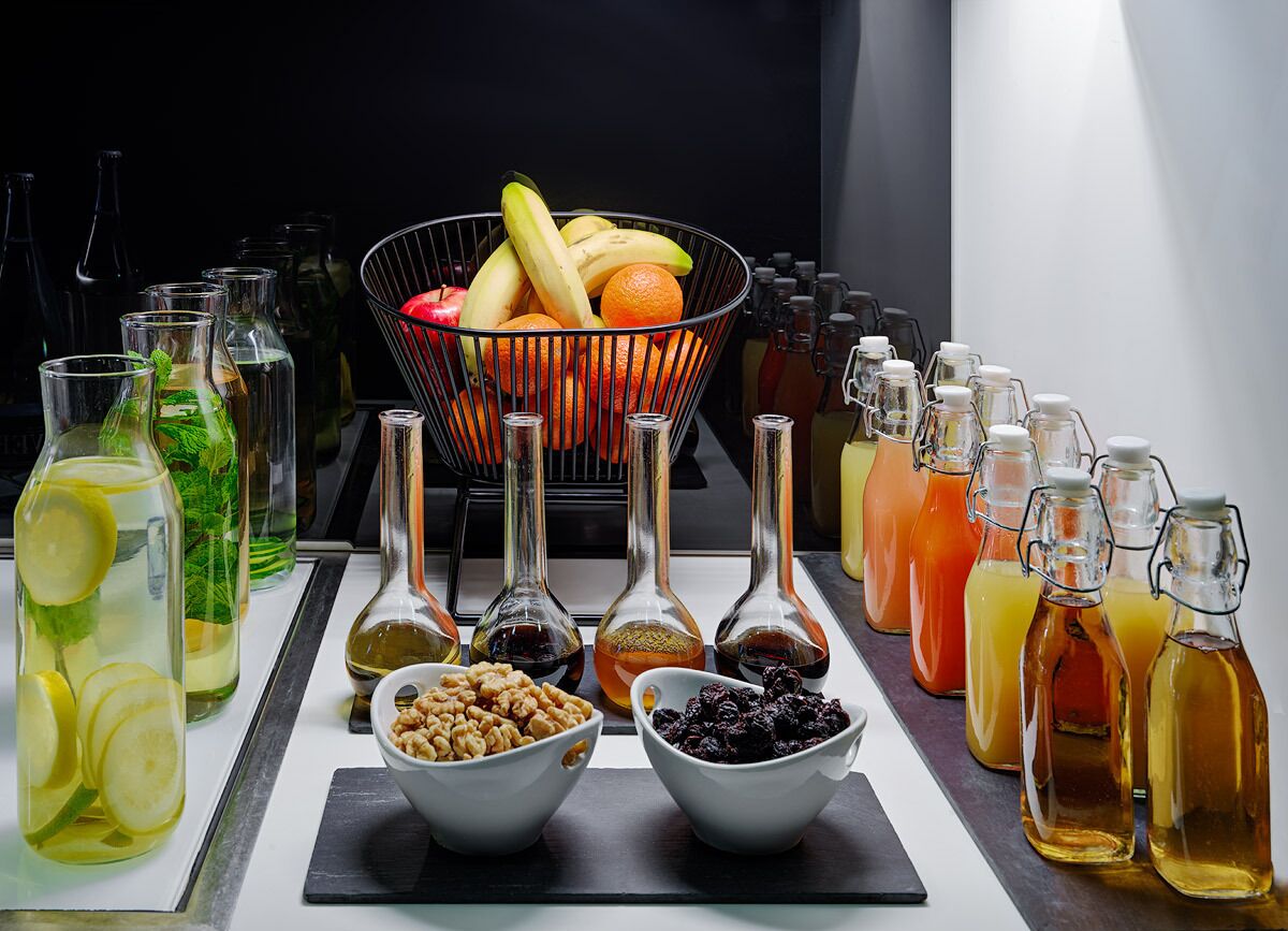Variedad de bebidas y frutas sobre una mesa, incluyendo botellas con diferentes líquidos y un bol con frutas mezcladas.