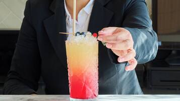 A person in a black blazer sitting at a marble table with a tall glass of a layered drink, a straw, and a cherry on top. Behind them is a bar with various bottles and glasses.
