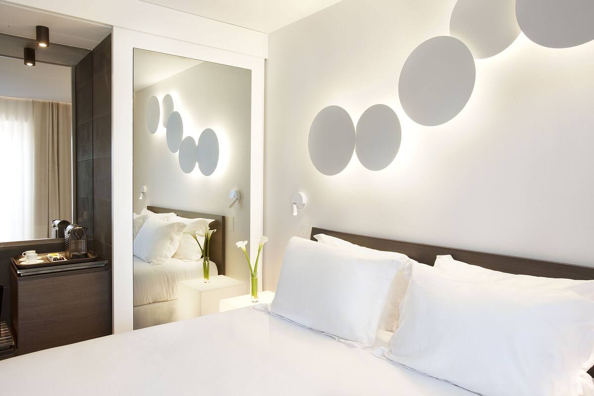 A modern hotel room with a neatly made bed, a bedside table with a lamp, and a mirror reflecting the room's decor.
