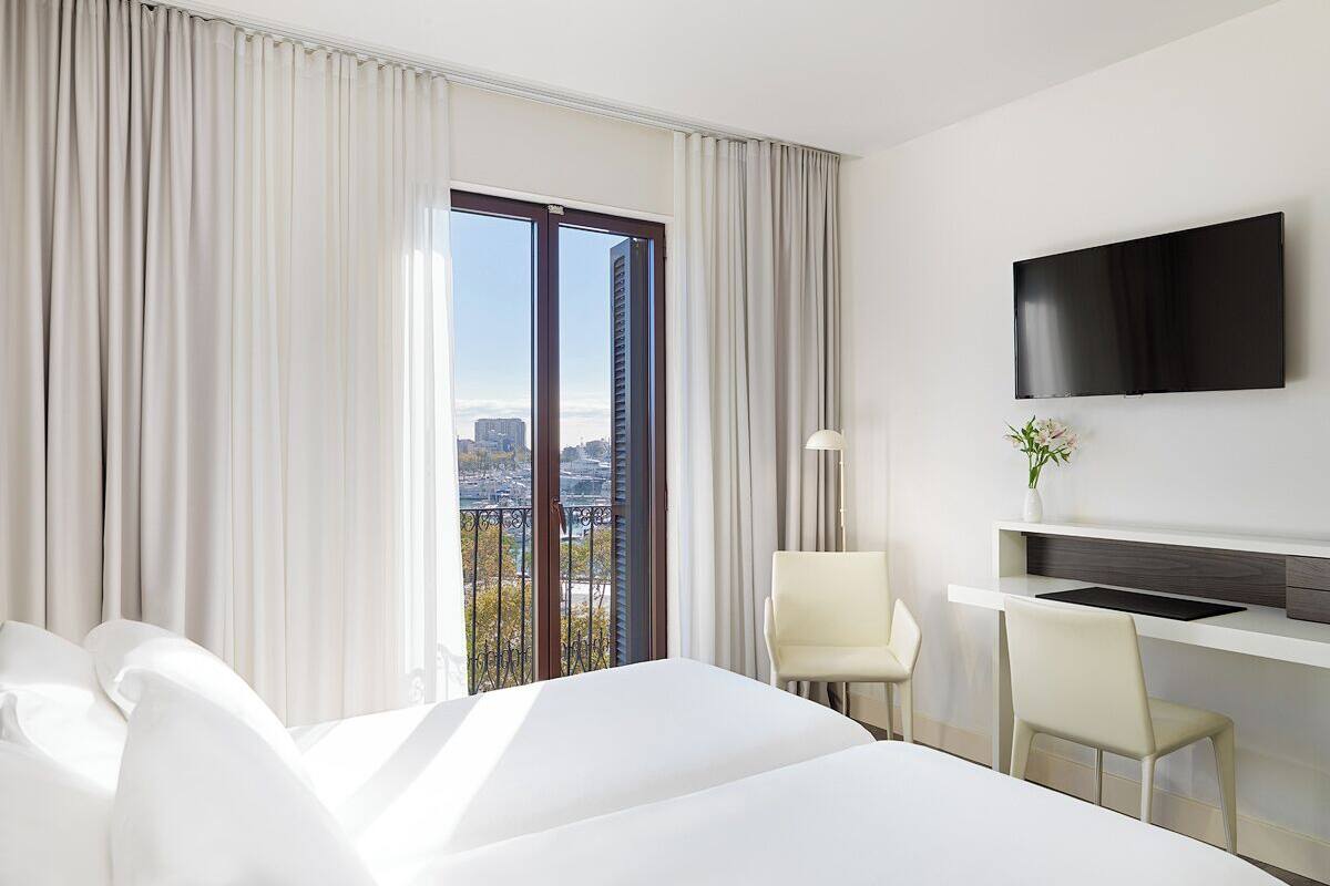 Habitación de hotel con una cama grande, un escritorio, un televisor de pantalla plana, una silla y vistas de la ciudad a través de un ventanal.