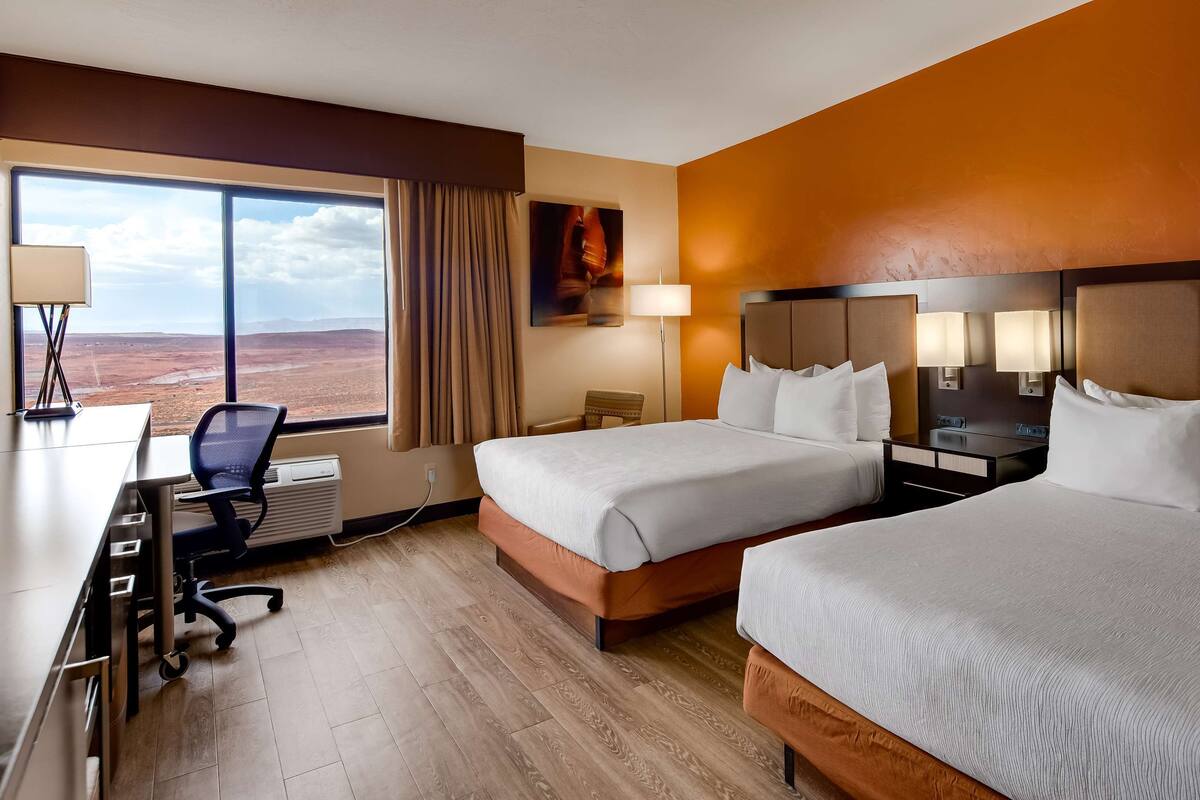 A hotel room with two beds, a desk, a chair, and a large window offering a desert view.
