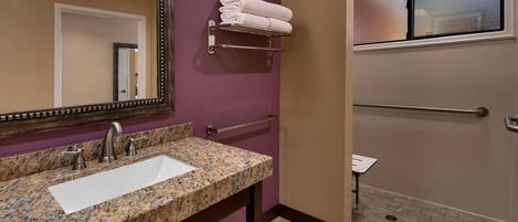 A bathroom with a granite countertop sink, a mirror with a decorative frame, a towel rack with folded towels, a handrail, and a window with frosted glass.