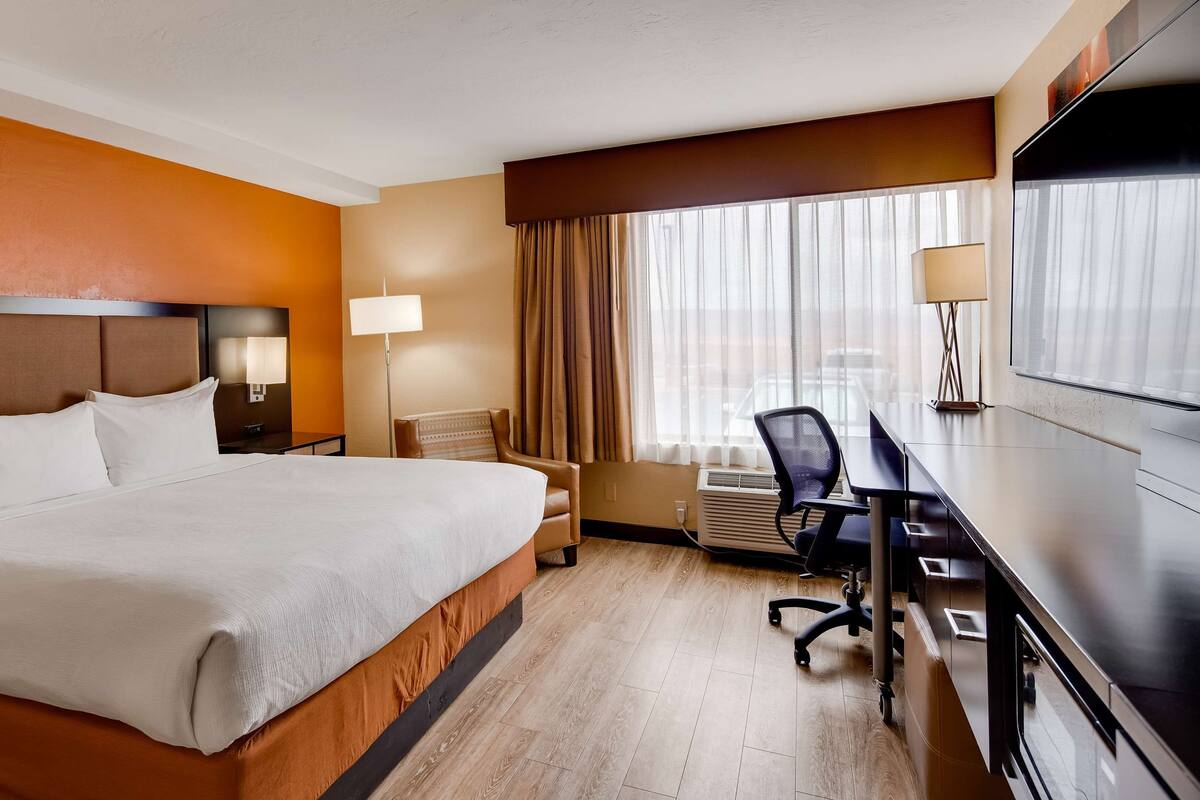 A hotel room with a large bed, a desk with a chair, a lamp, and a window with curtains.