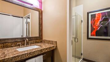 A bathroom with a granite countertop, a sink, a mirror, a glass shower door, and a framed artwork on the wall.