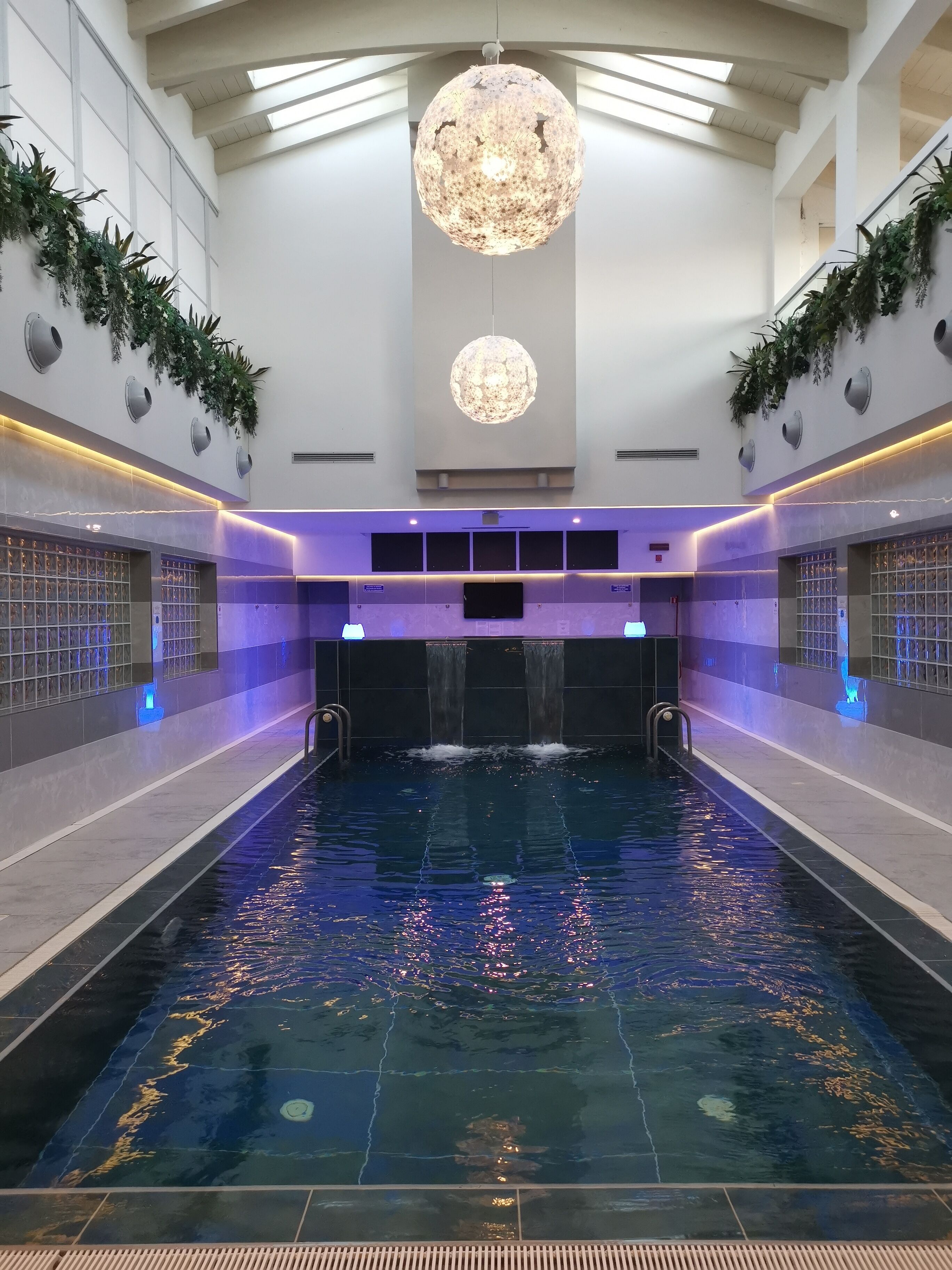 Une piscine intérieure dotée d’une cascade centrale, entourée de murs agrémentés de jardinières intégrées et surmontée d’un lustre moderne.