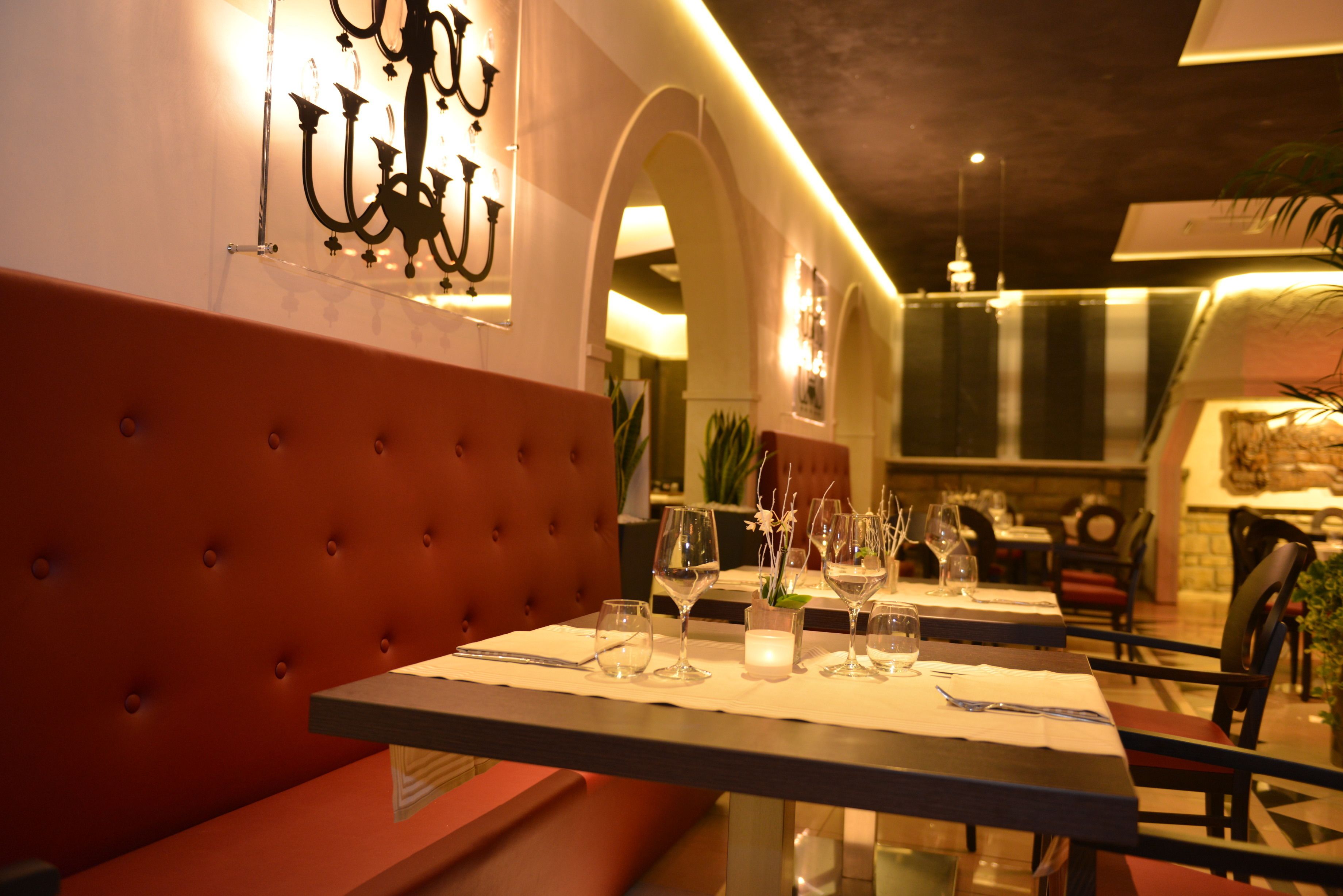 A dining area with a table set for two, featuring a tufted red upholstered bench, a chandelier, and a view of the restaurant's interior.