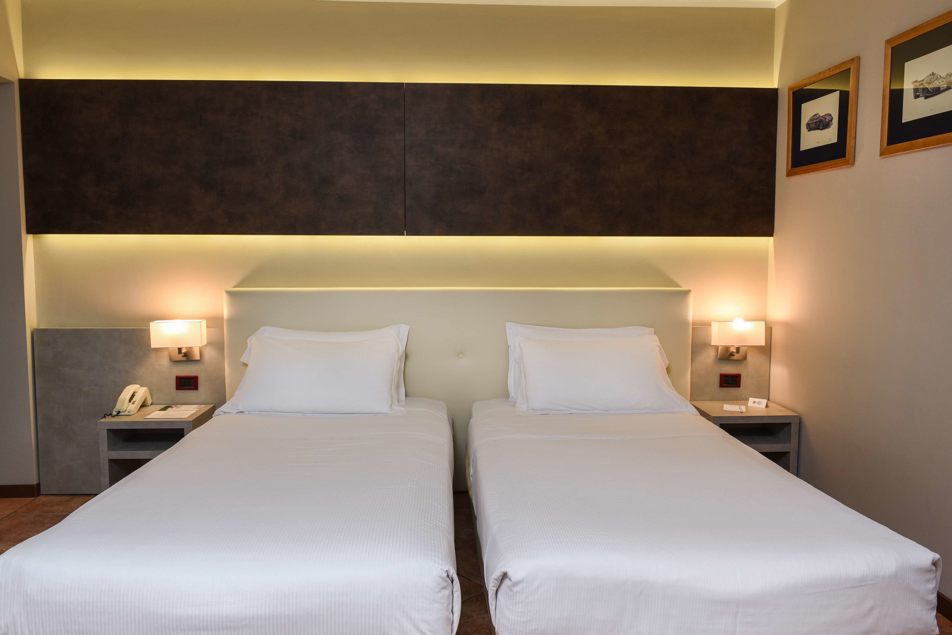 A hotel room with two single beds, a large headboard, bedside lamps, a phone, and framed pictures on the wall.