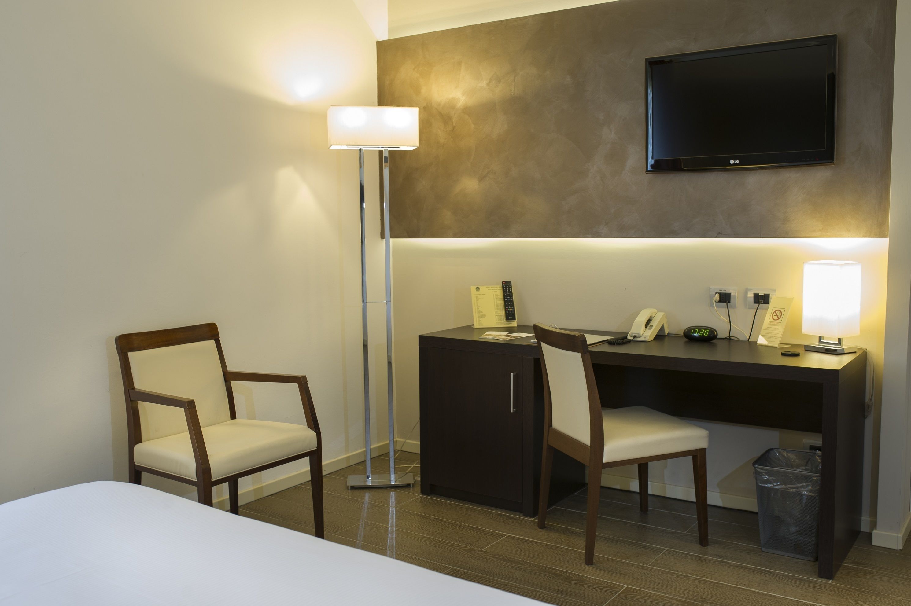 Hotelkamer met een bed, een bureau met stoel, bureaulamp, telefoon, televisie aan de muur en een prullenbak.