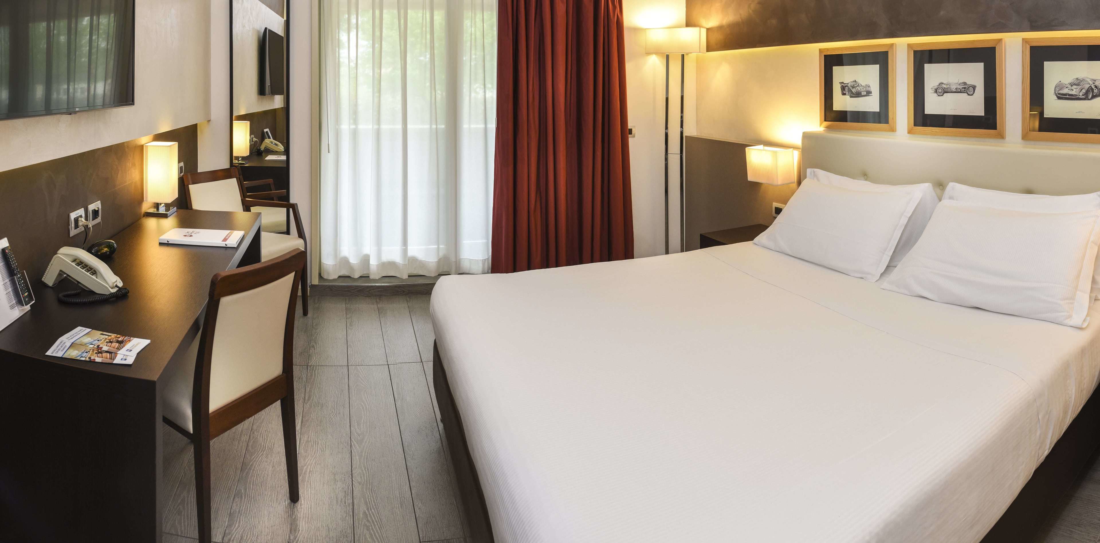 A hotel room with a large bed, a desk with a phone, a chair, a window with curtains, and framed pictures on the wall.