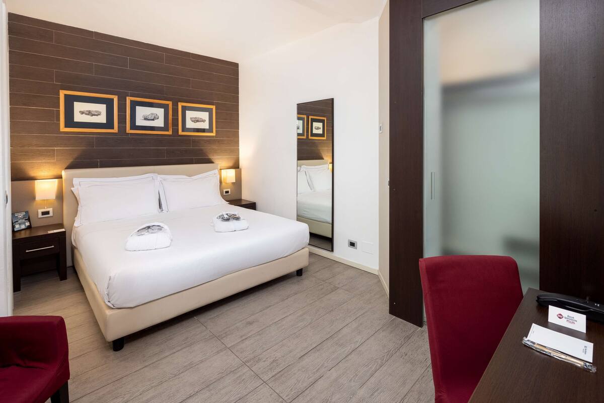 A hotel room with a bed, two bedside tables, a mirror, a red chair, and a wooden floor.