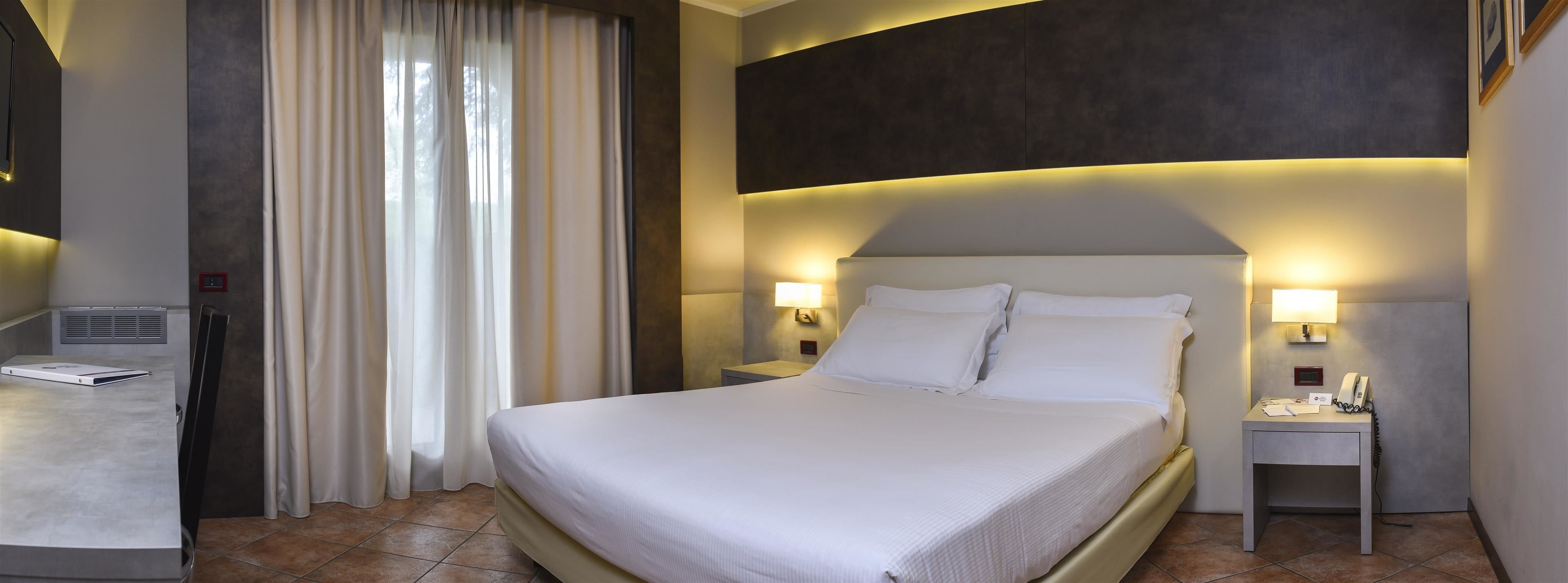 Quarto de hotel com uma cama grande, duas luminárias de cabeceira, uma escrivaninha com computador e uma janela com cortinas.