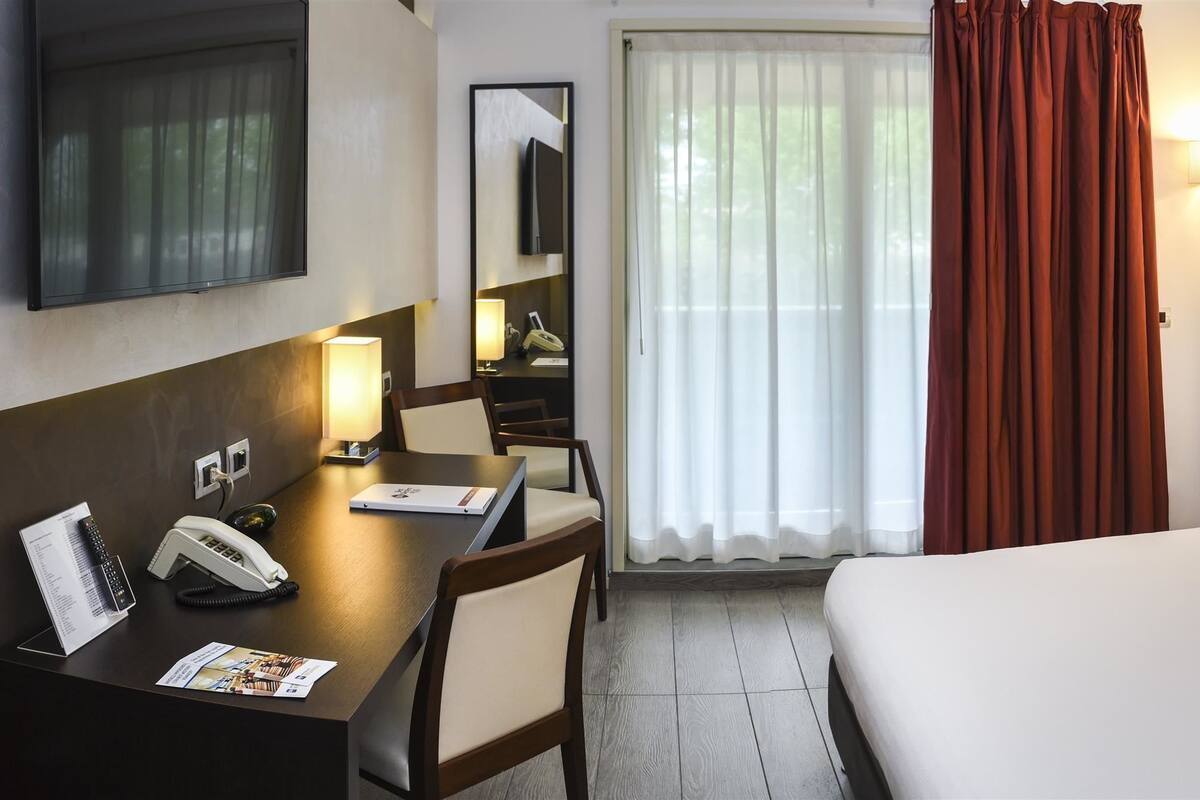 A hotel room with a bed, a desk with a telephone, a mirror, and framed pictures on the wall.