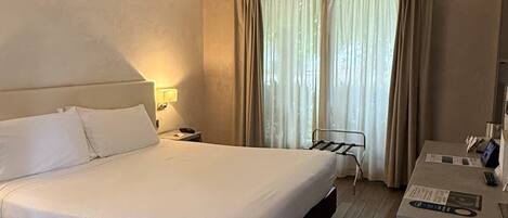 Quarto de hotel com cama grande, mesa de cabeceira, abajur e escrivaninha com cadeira.