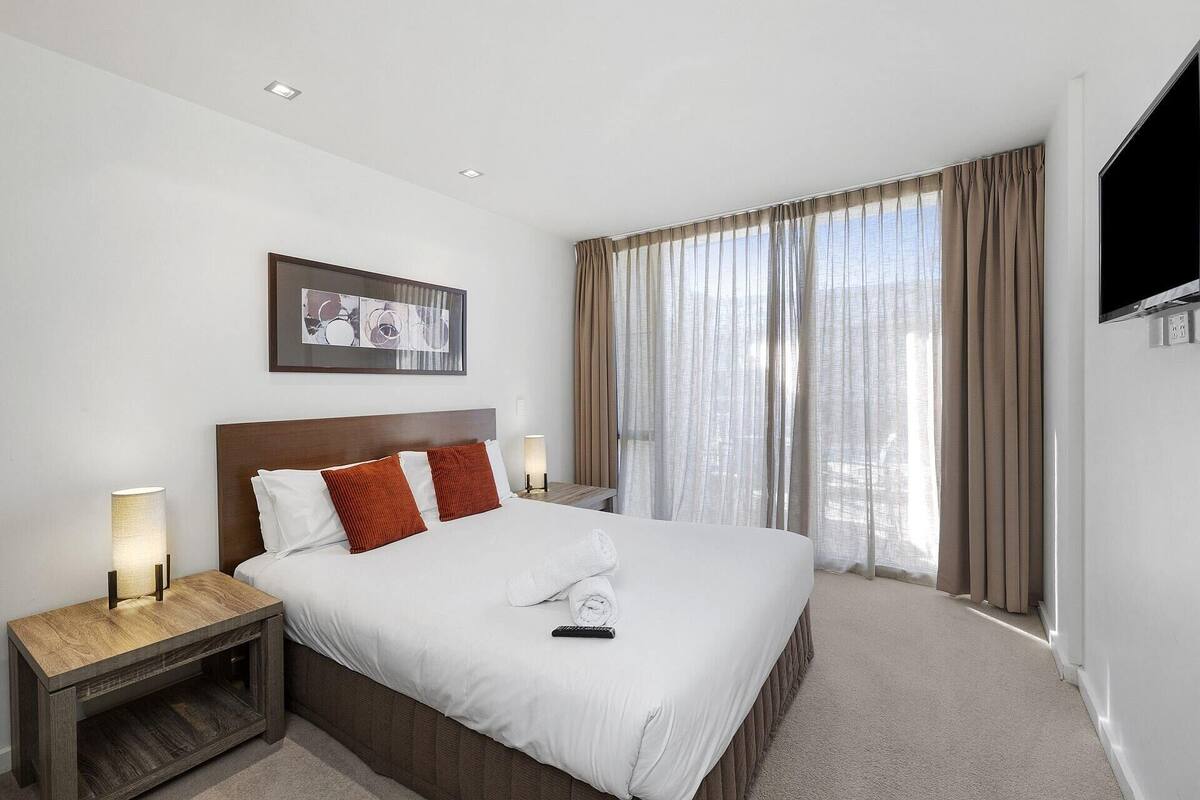 Une chambre d’hôtel avec un grand lit, une tête de lit, deux tables de chevet avec des lampes, un téléviseur à écran plat fixé au mur et une fenêtre avec des rideaux.