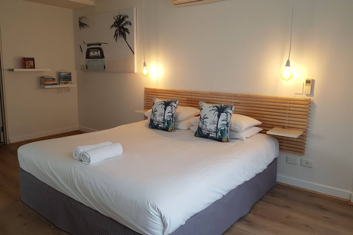 Un lit bien fait, avec du linge de lit blanc et deux coussins décoratifs, une table de chevet avec une lampe et une étagère avec des livres et un petit objet décoratif.