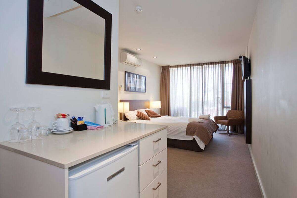 Une chambre d’hôtel comprenant un lit, un bureau avec des tiroirs, un miroir, une télévision et une fenêtre avec des rideaux.