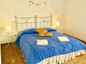 Cama com colcha azul, duas almofadas e duas toalhas brancas. Há duas mesas de cabeceira com luminárias.
