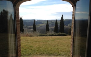 A view through an arched window of a grassy field with several tall cypress trees and rolling hills in the distance. - Agriturismo Podere Fornaci (Asciano)