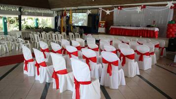 A hall with rows of white chairs with red sashes, a stage with a red tablecloth, and a screen displaying an image.